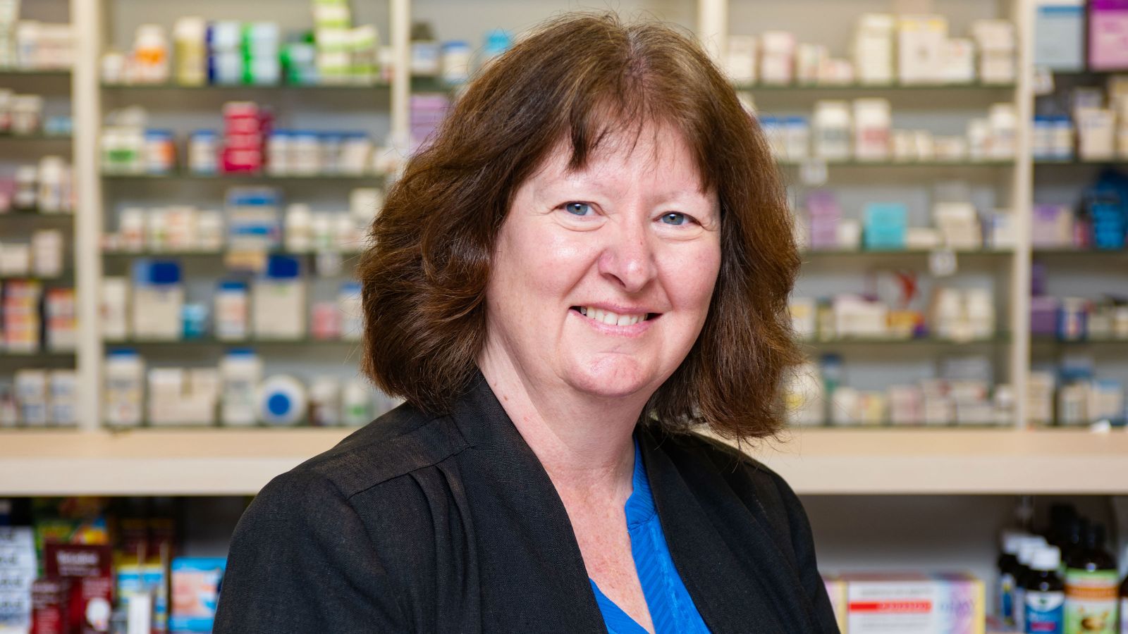 Professor Jacqueline Cumming stands and smiles before a mosaic of pharmaceutical bottles and boxes.