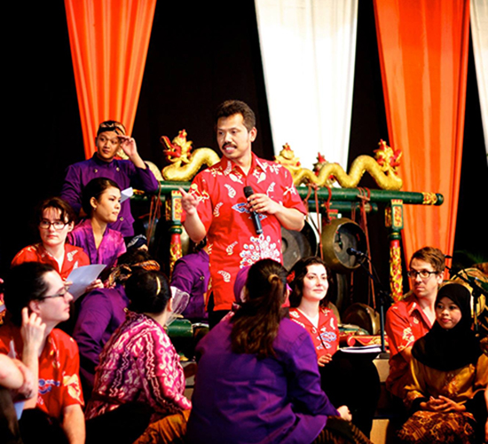 A male Indonesian gamelan music teacher talks to a group of students dressed in red and gold.