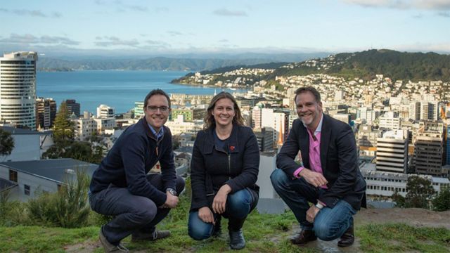 Two men and a woman kneel down for a group photo with Wellington central city behind them.