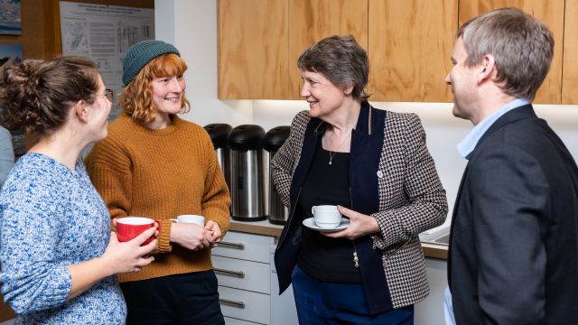 Helen Clark and three others stand next to a kitchenette, chatting and holding coffee cups.  