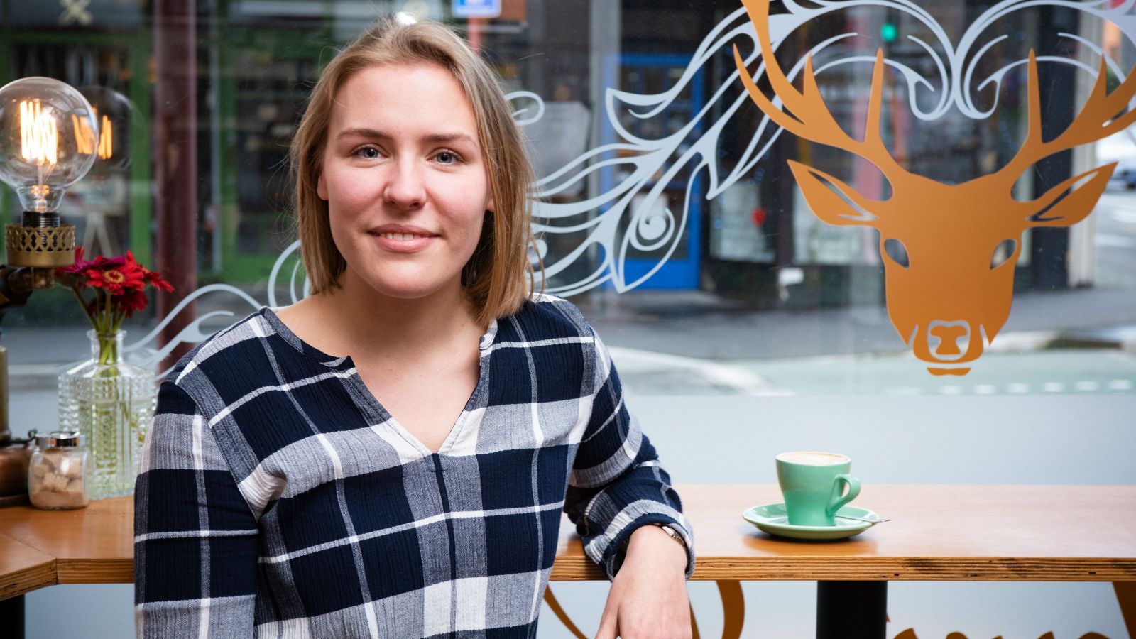 Yvonne looks straight at the camera. She sits by the window in a cafe with a green coffee mug next to her on the bench. 