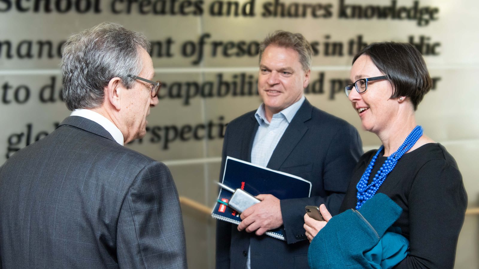 A man in a suit and a woman wearing a blue necklace talk with Professor Girol Karacaoglu at Victoria Business School during an event.