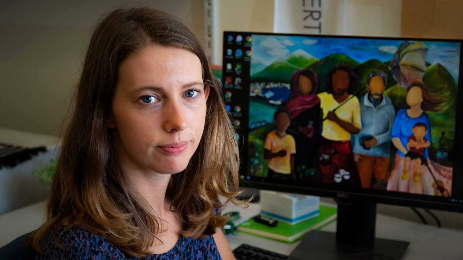 Amber sits at her desk and looks directly at the camera. Behind her, on her computer screen, is one of the paintings from her exhibition.