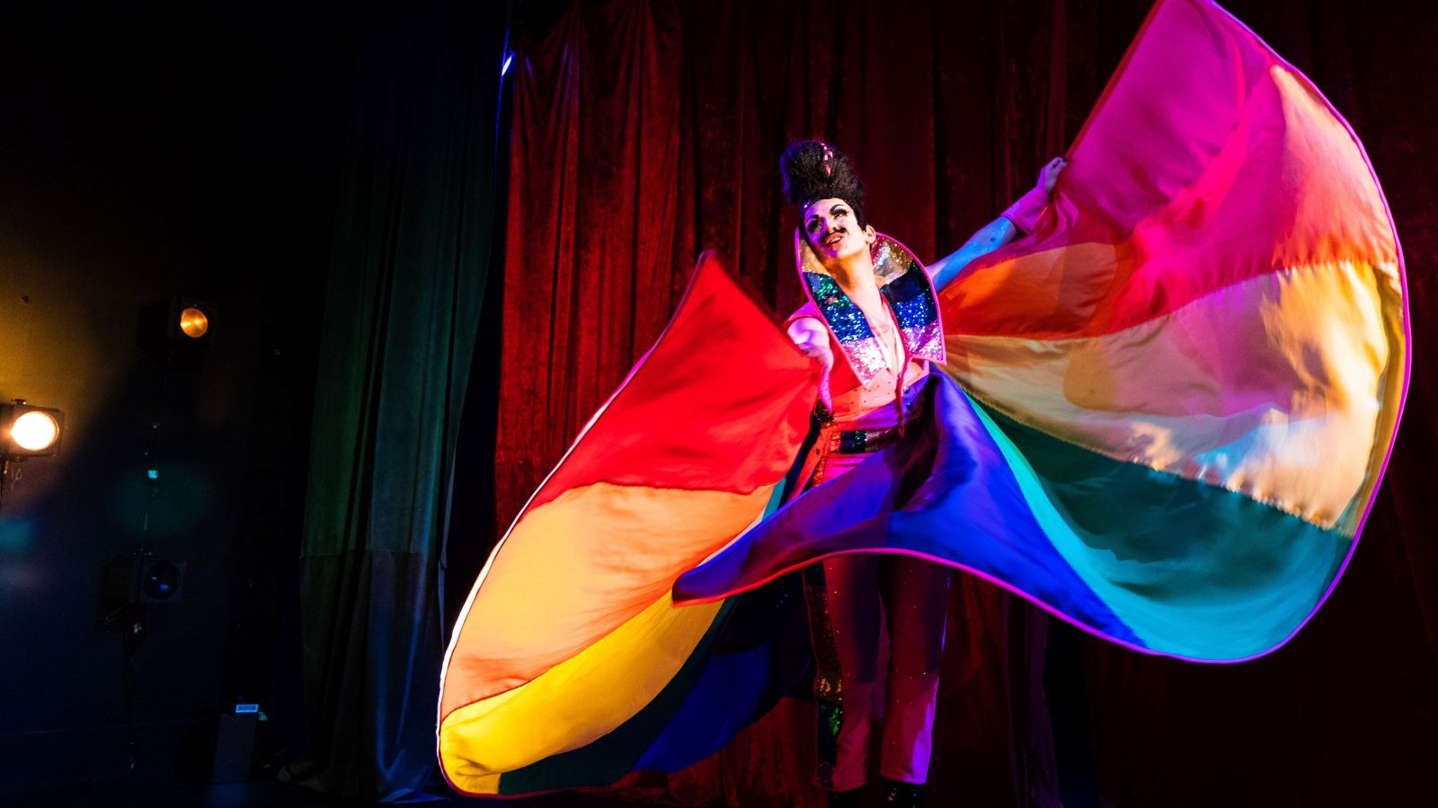 Drag King dances in a pink jumpsuit with a rainbow cape.