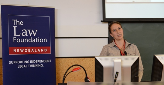 A professional woman speaks at the front of a lecture hall with the a poster in the background that reads, The Law Foundation of New Zealand, Supporting Independent Legal Thinking.