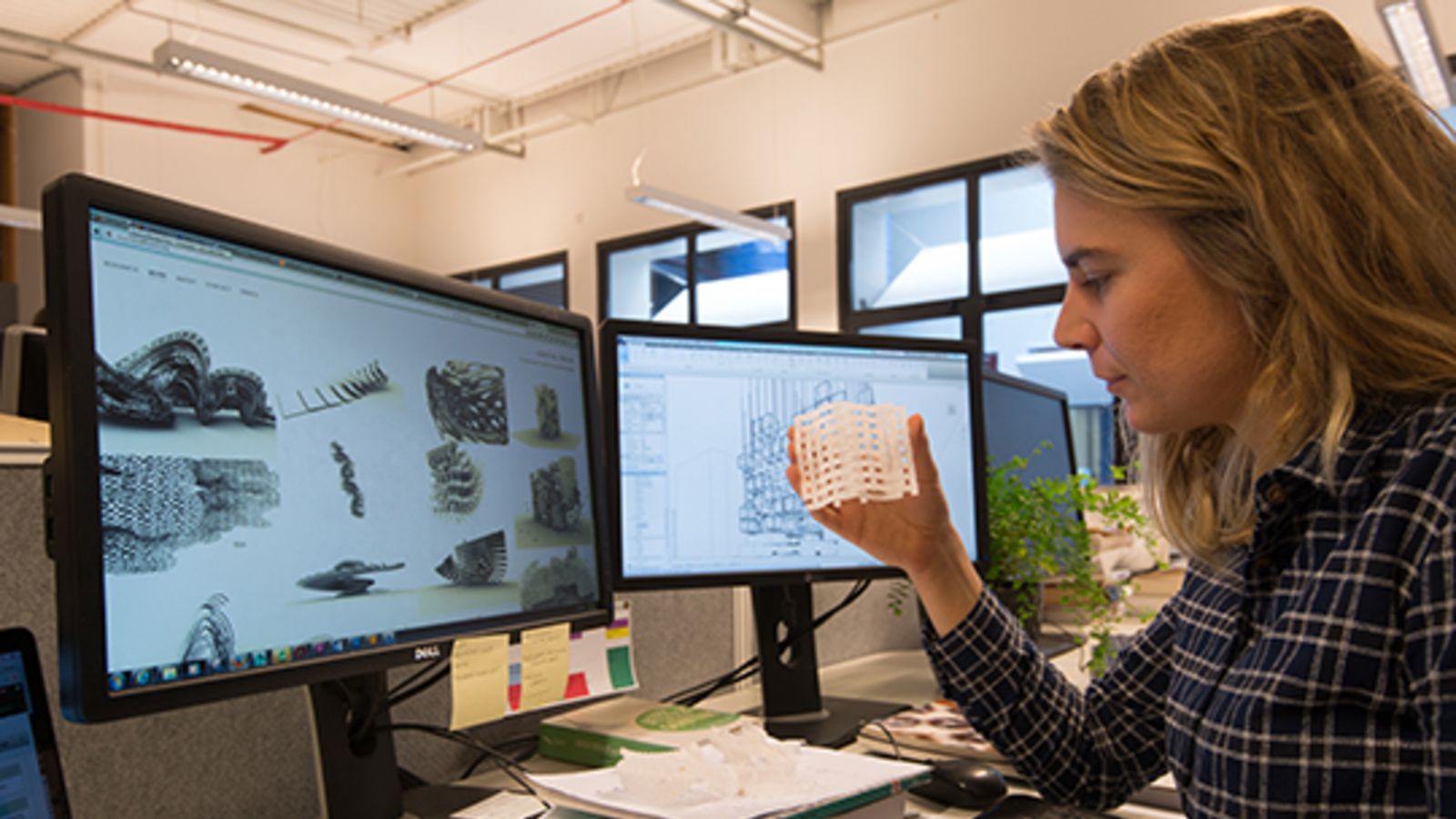 Claudia van Velthooven holding a 3D printed object in front of two computer screens.