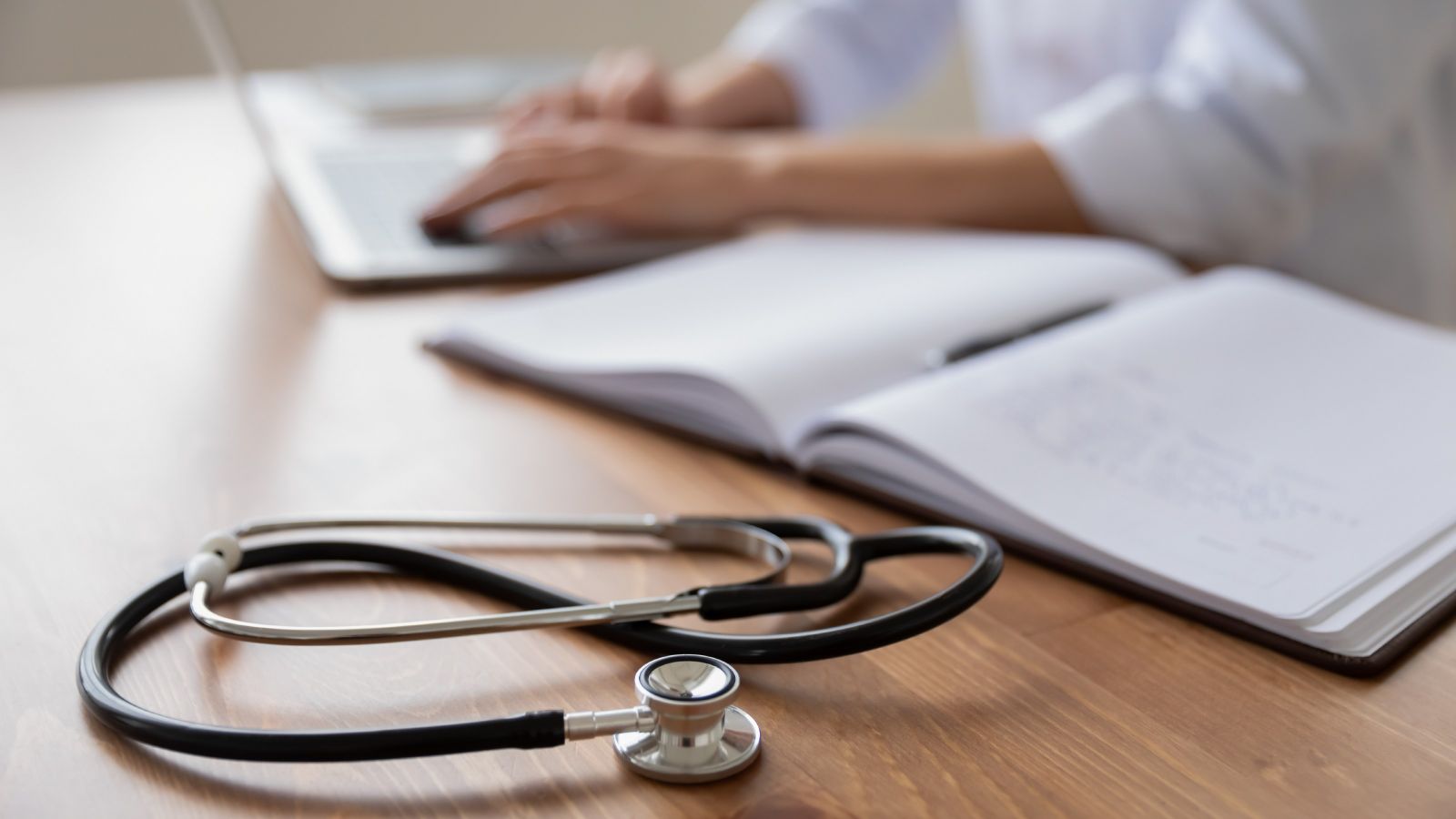 A stethoscope in the foreground of a desk where a general practitioner sits at a laptop computer