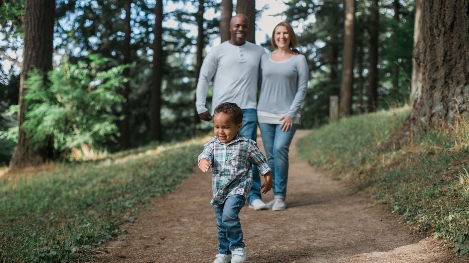 dark skinned baby running ahead in forest, with dark skinned dad and white mum