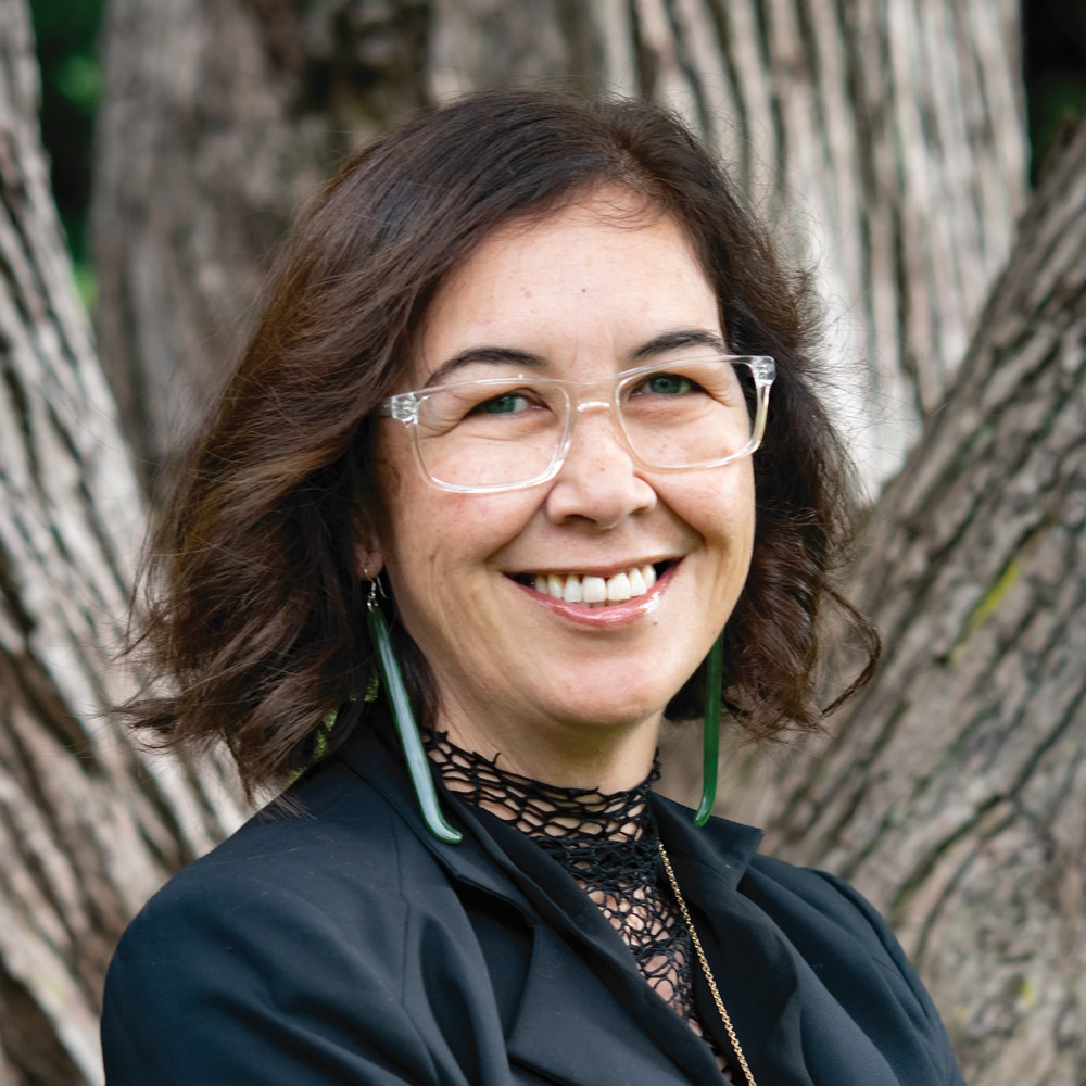 Rawinia Higgins, a smartly dressed Māori woman in front of tree trunks.