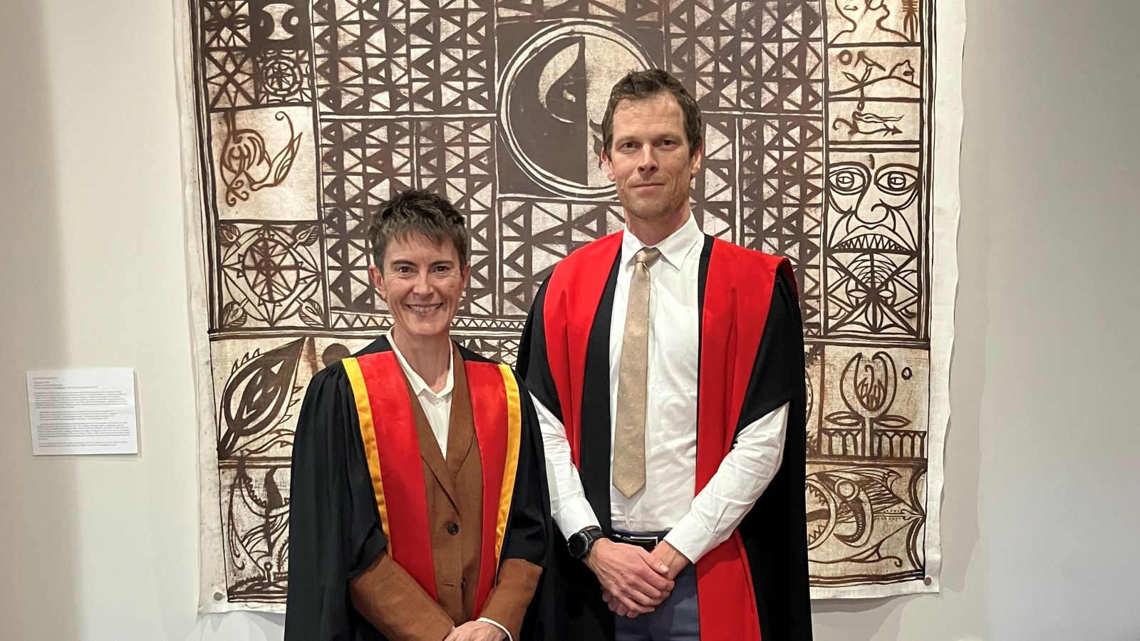 Two professionally dressed academics with gowns standing in front of a piece of artwork.