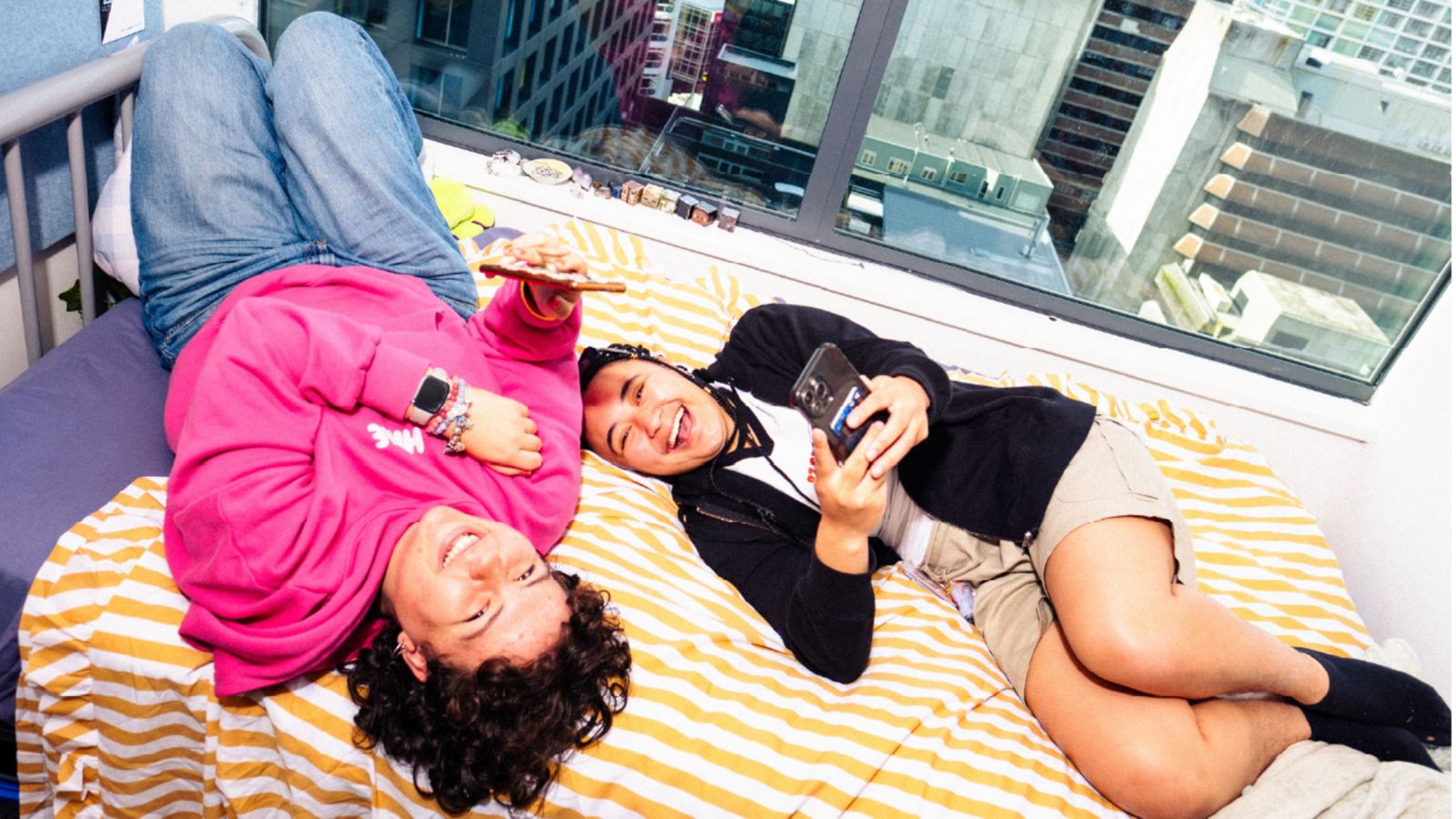 Two smiling people lying on a bed with striped bedding, each holding a phone, in a room with large windows showing city buildings.