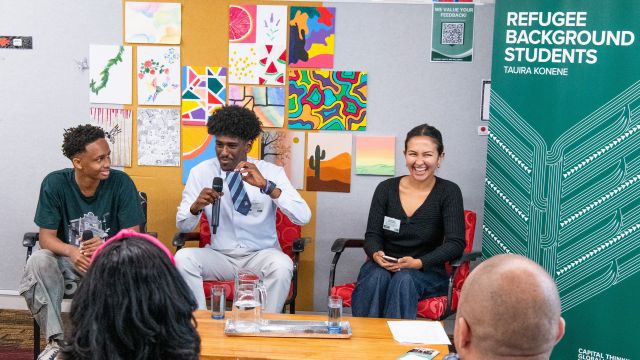 A student panel featuring two students and a moderator smiling in The Bubble next to the Refugee-background students banner.