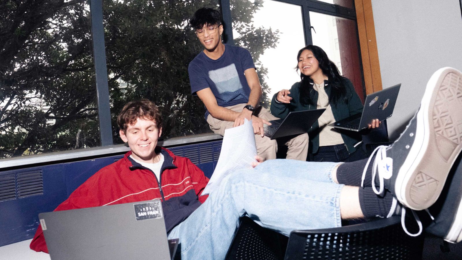 3 students near a window look at a laptop of the male student in the foreground.