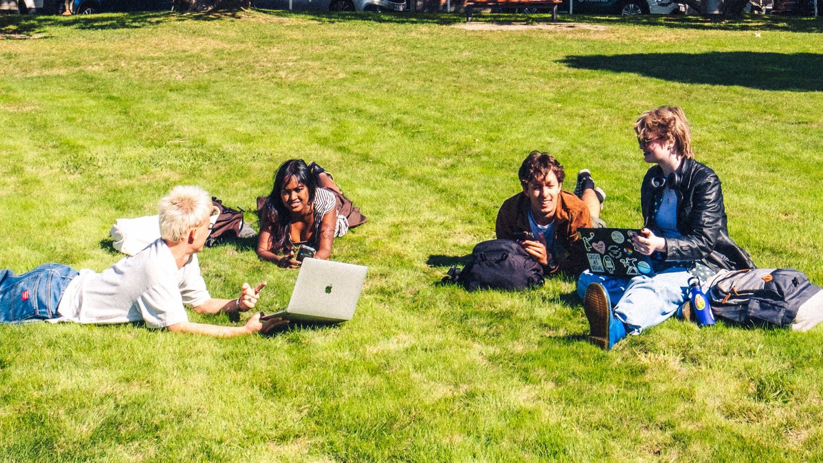 four students lying on the grass outside a university building studying on laptops
