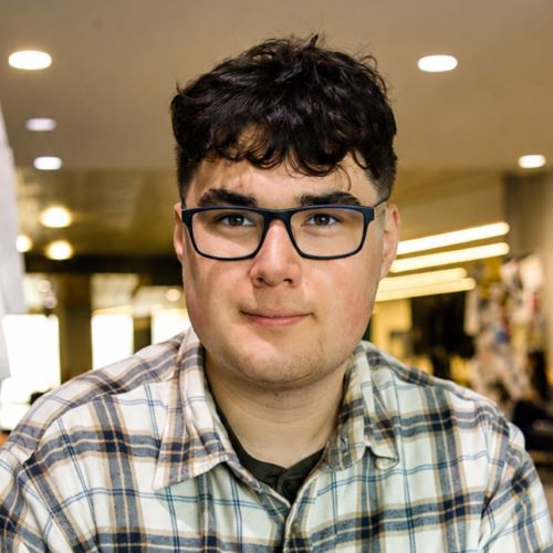 Person in glasses smiling in a large room, wearing a plaid shirt. 