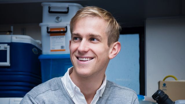 A profile image of Evan Brenton-Rule looking off past the camera, smiling. Behind him are storage boxes and the top of a microscope.