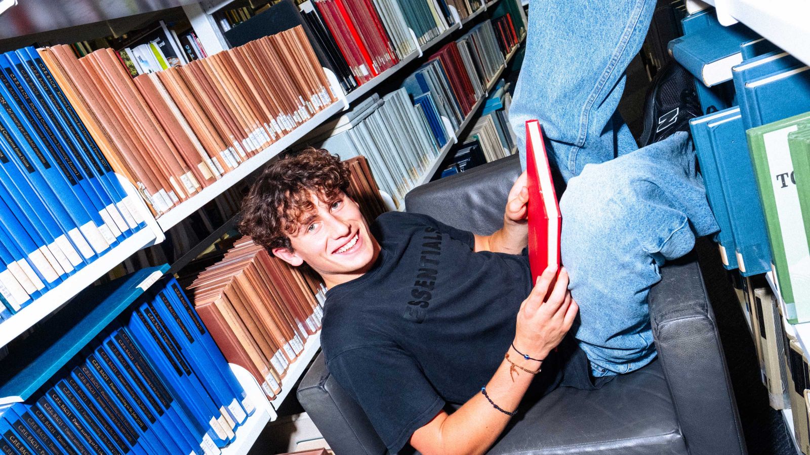 Student slouching sideways on an armchair in the library, in between shelves of books, with a book with a red cover on their lap.