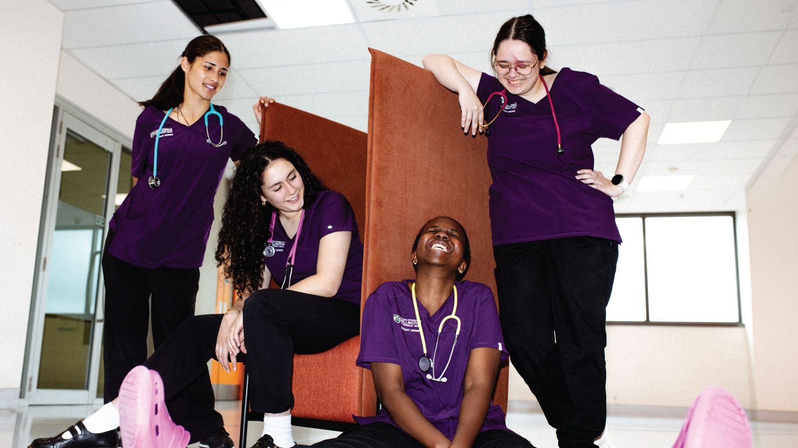 Four female midwifery students hanging out in a room, smiling and laughing together wearing scrubs.