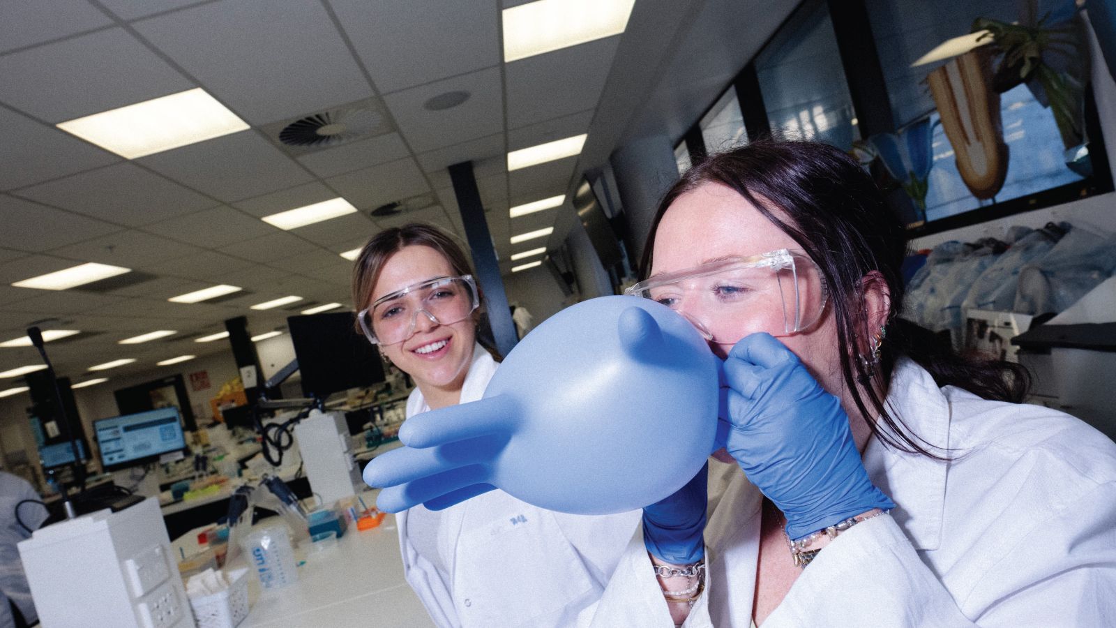 Two science students wearing lab coats and safety glasses, smiling and blowing into a rubber glove