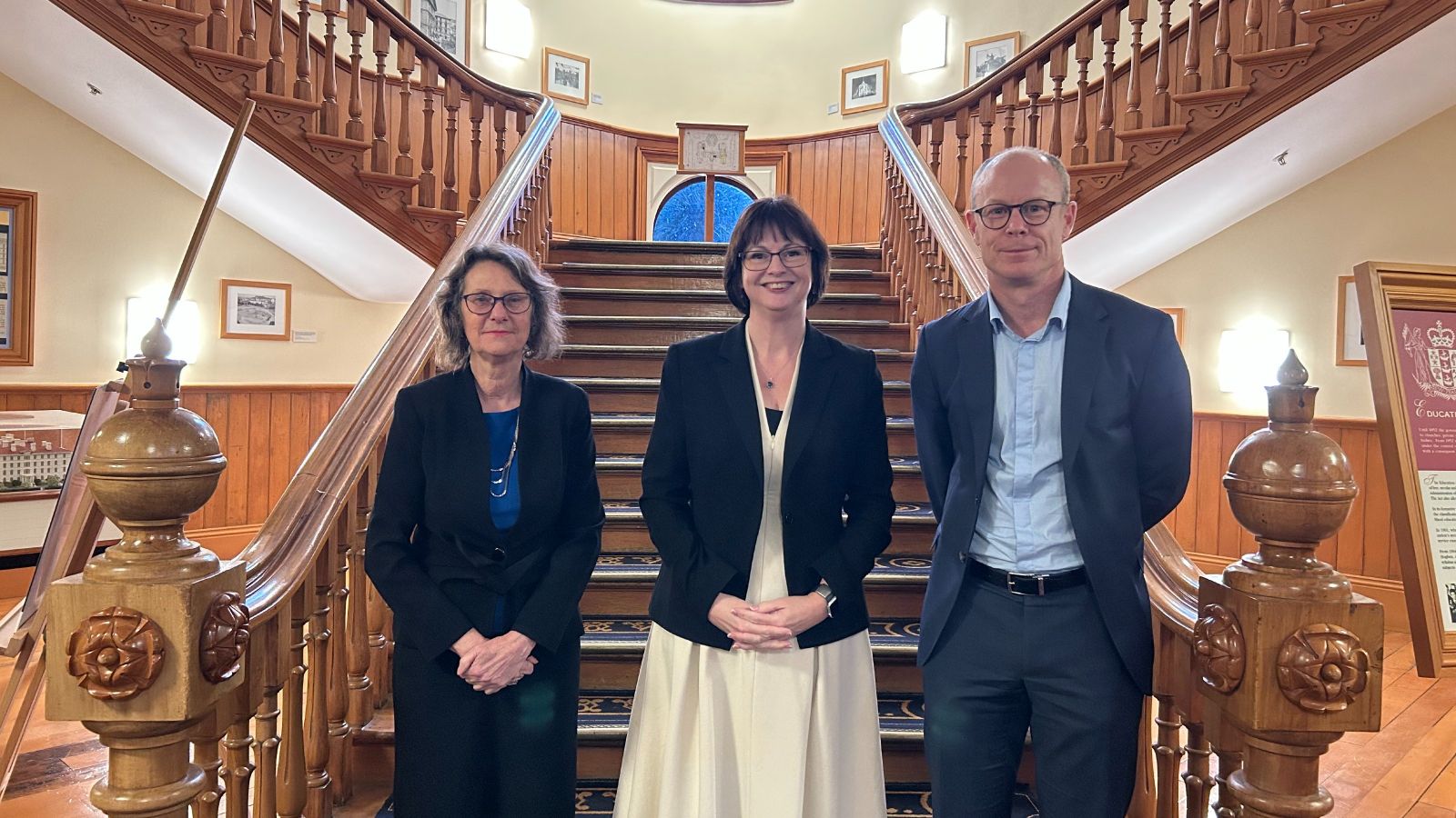 Three professionally dressed academics standing in front of a wooden staircase.