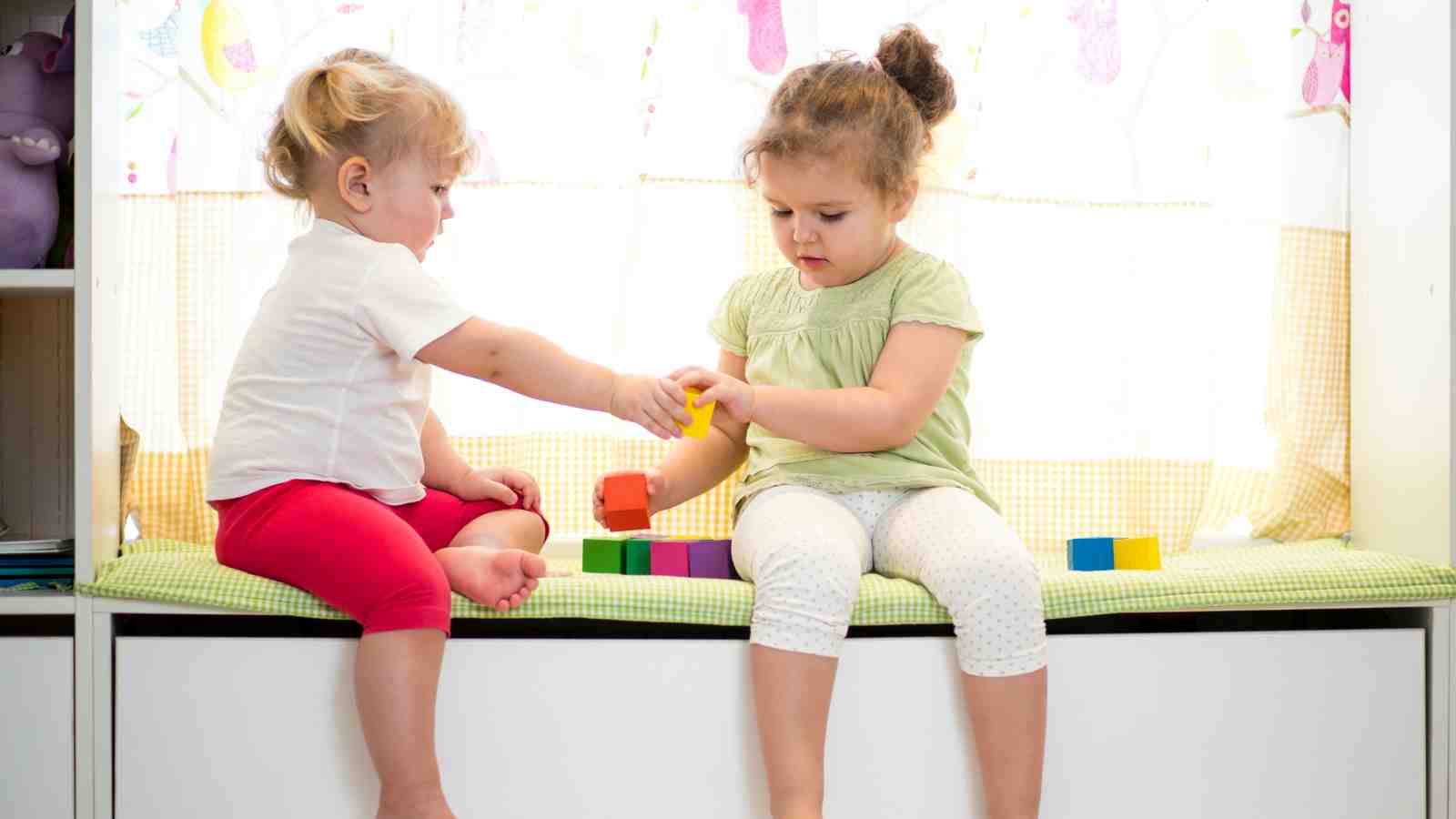 two girls playing with coloured blocks while seated on a checkered green bench.