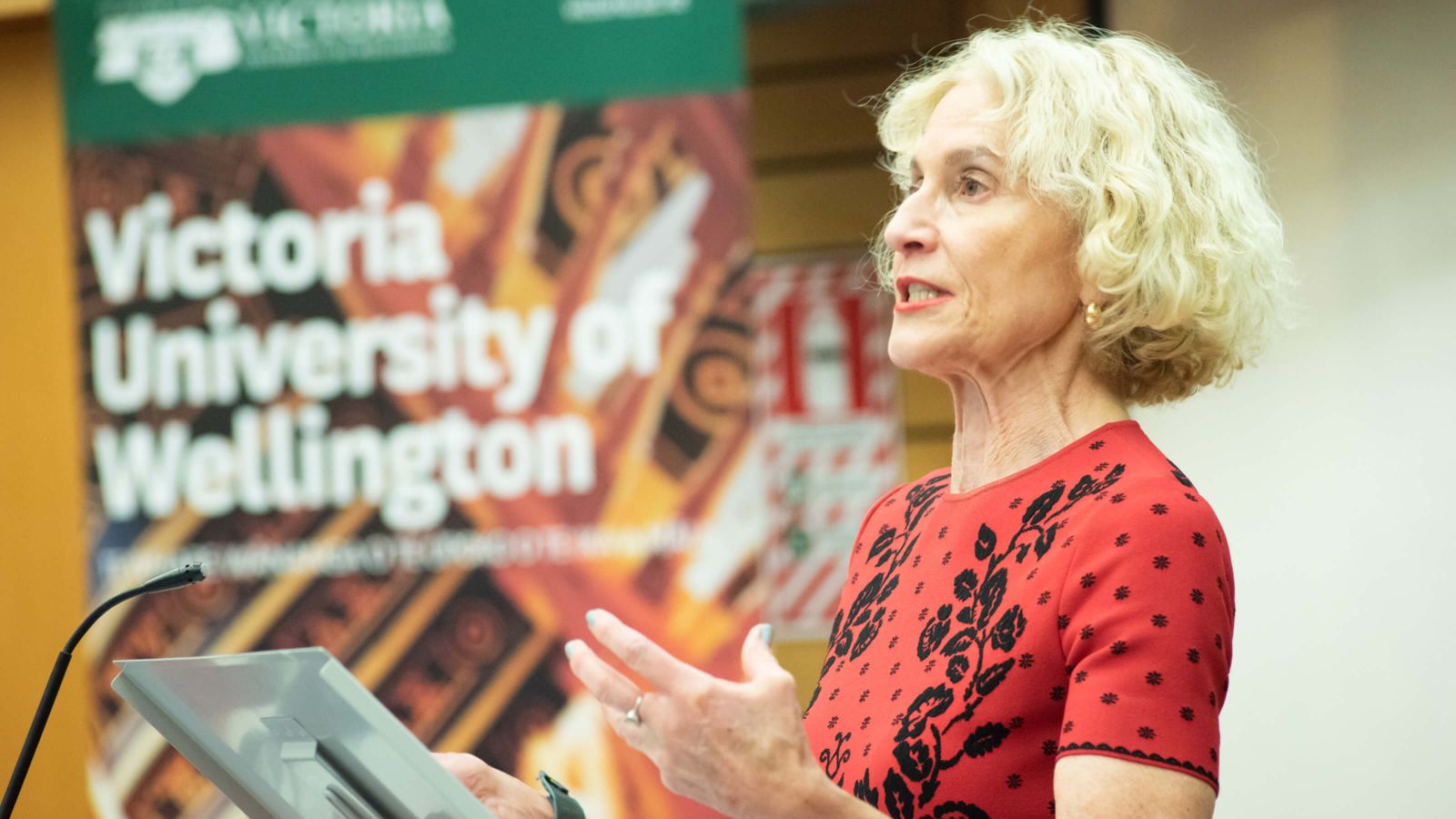 Blonde woman in red clothes speaks over a metal lectern. 