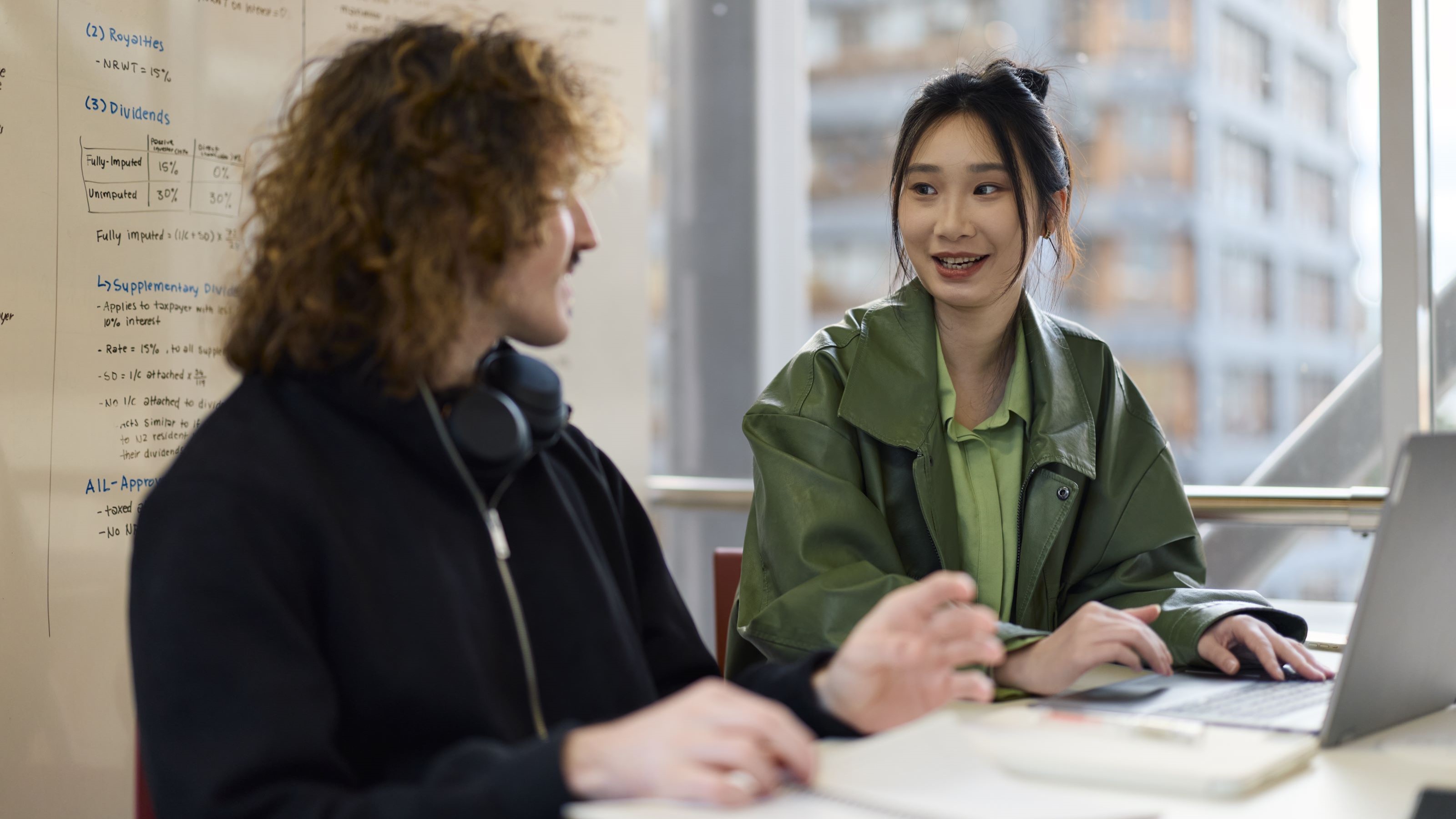 2 students sitting in front of a laptop, with a whiteboard behind them. They are in discussion