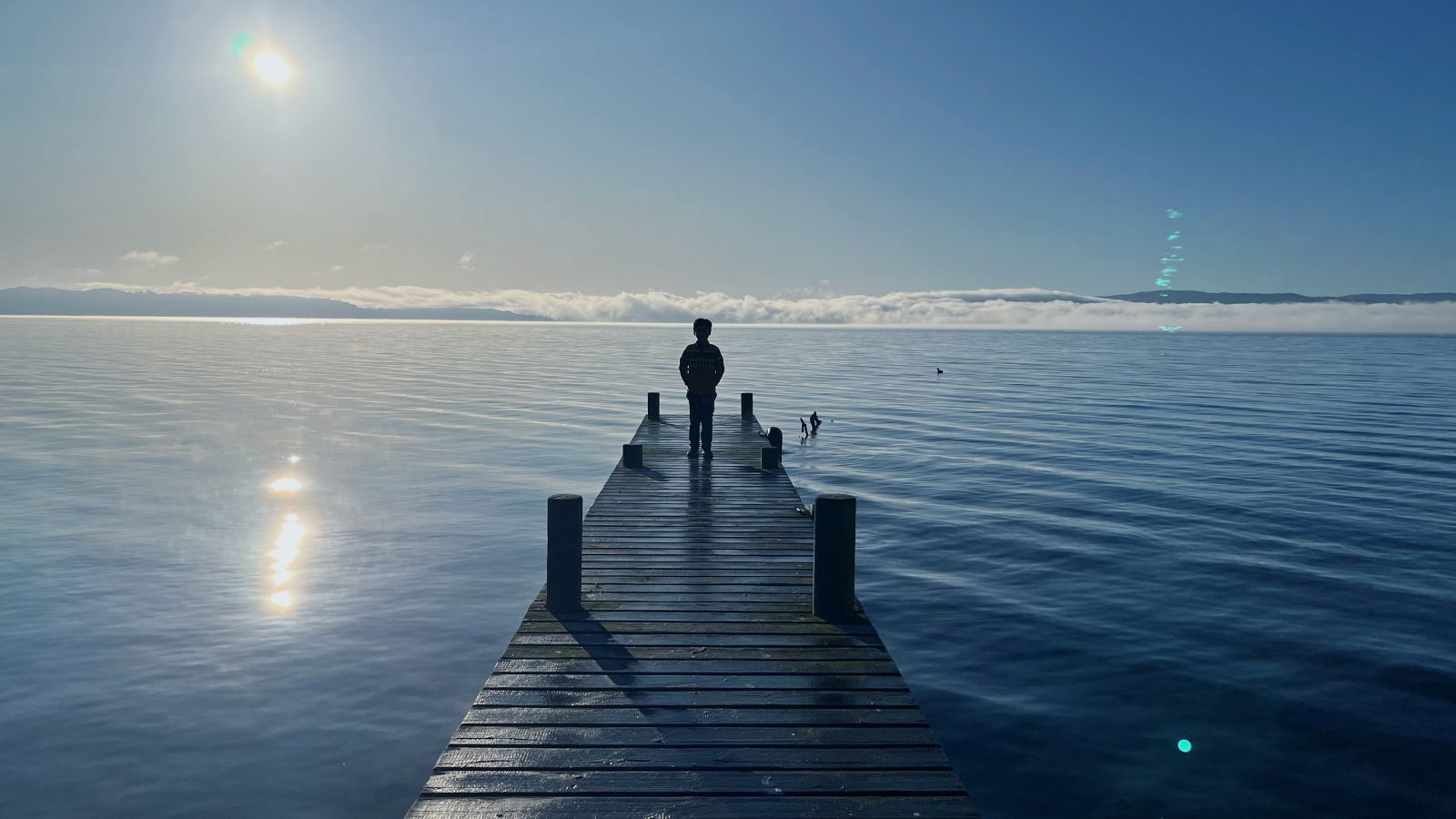 A child stands on the end of a pier looking out to a sparkling sea and clouds