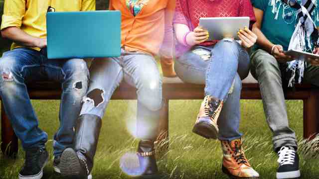 Mulicultural society – A group of students in colourful tops sit together on a bench with a laptop and a tablet.