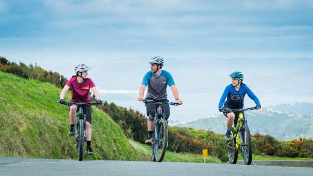 Three students on their mountain bikes, on a road, with mountain ranges and grass in the background.