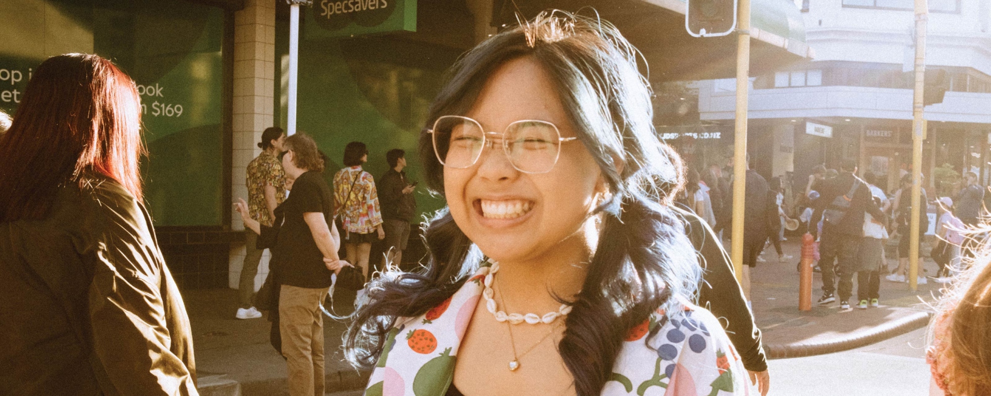 Street scene with several people walking, in the foreground, a student wearing a colourful fruit-patterned jacket, black top, and shell necklace is smiling.