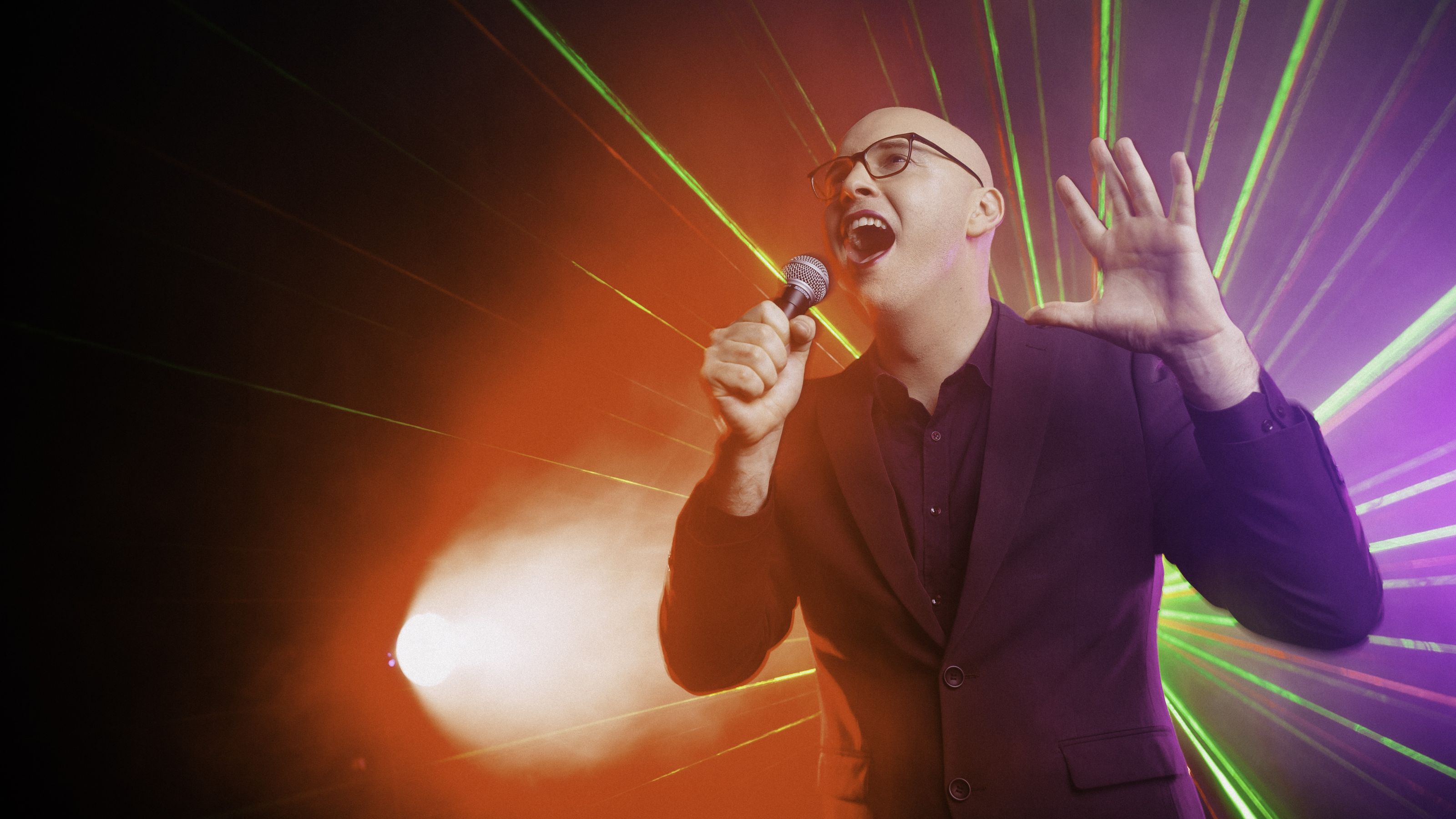 Dr James Wenley performing on stage with a microphone and light show in the background.