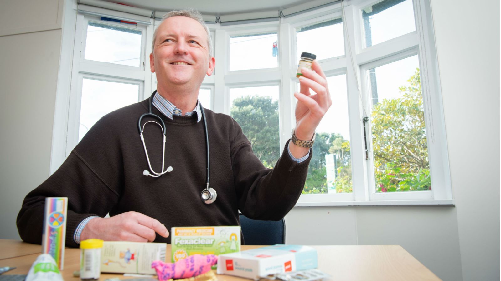 Professor Kevin Dew sits at a table, some common pharmaceuticals haphazardly spread before him, a white bay window in the rear.