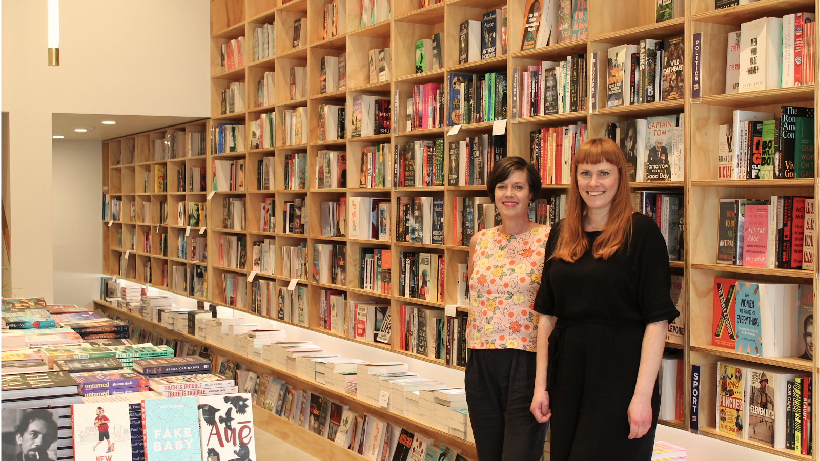 Two woman, one with long red hair, one with a brown bob, standing in front of a bookshelf