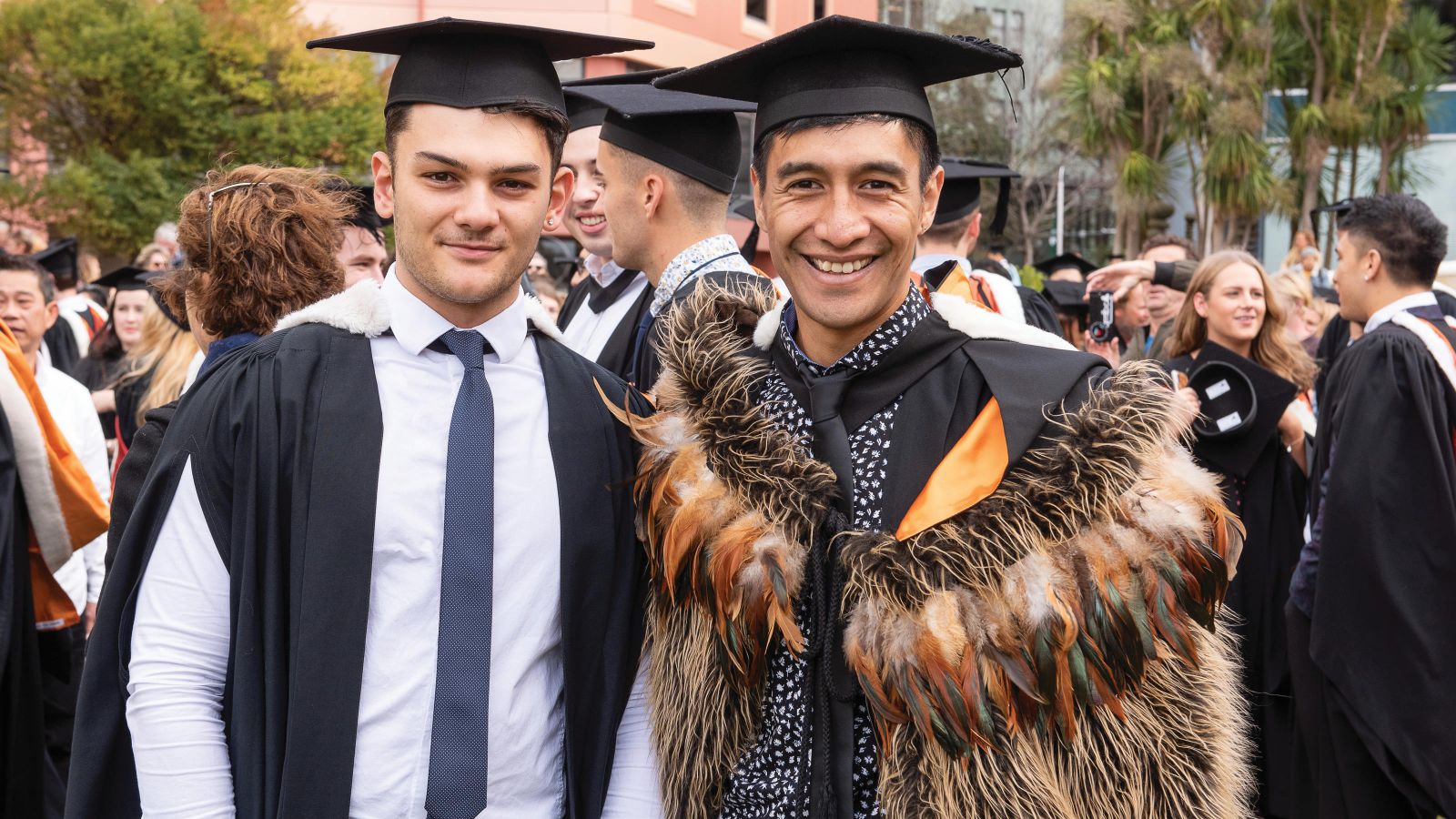 Two Māori men in graduation gowns, one also wearing a feather cloak, with more graduates in the background.