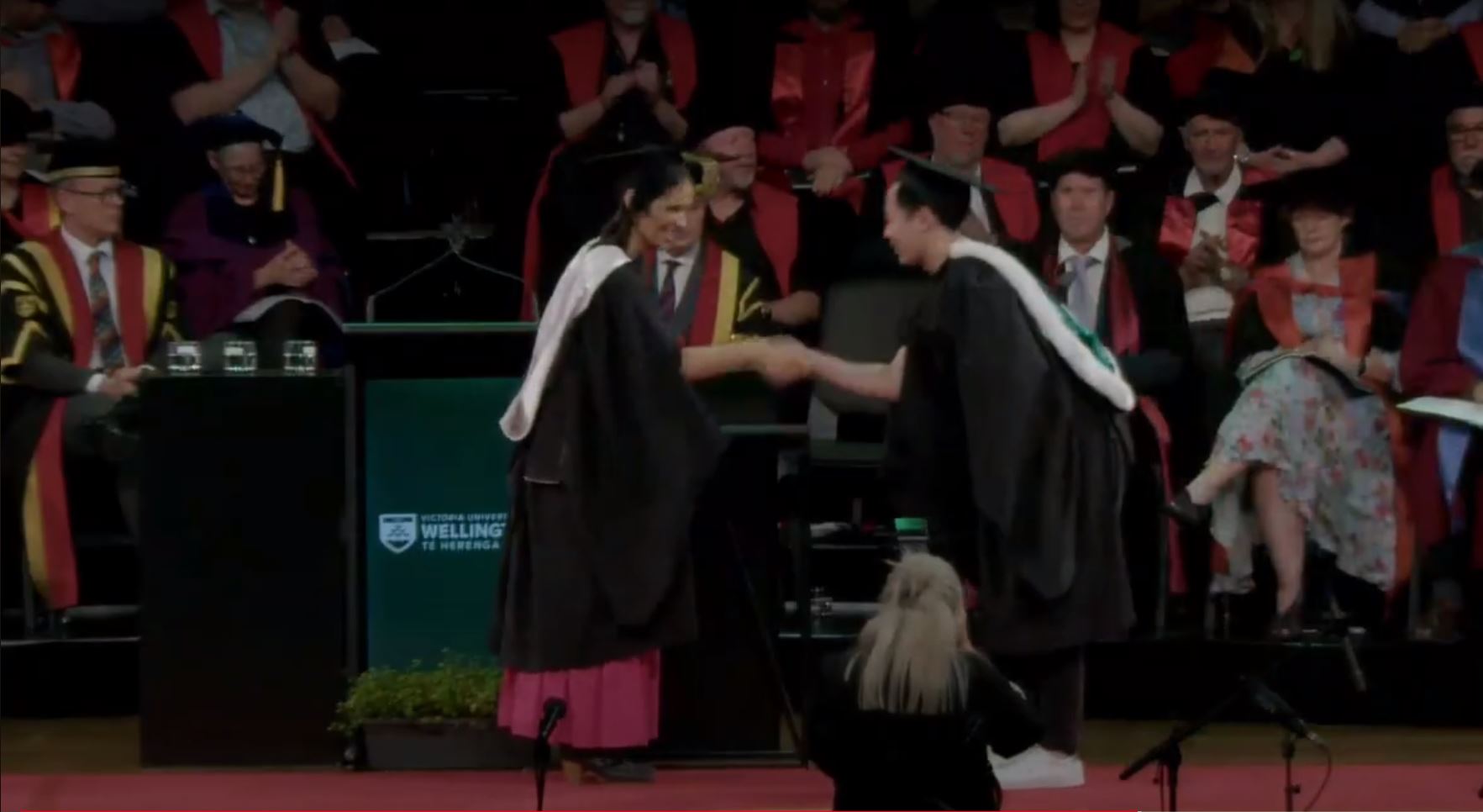 A graduate receiving their degree, with Nic Smith watching on the left.