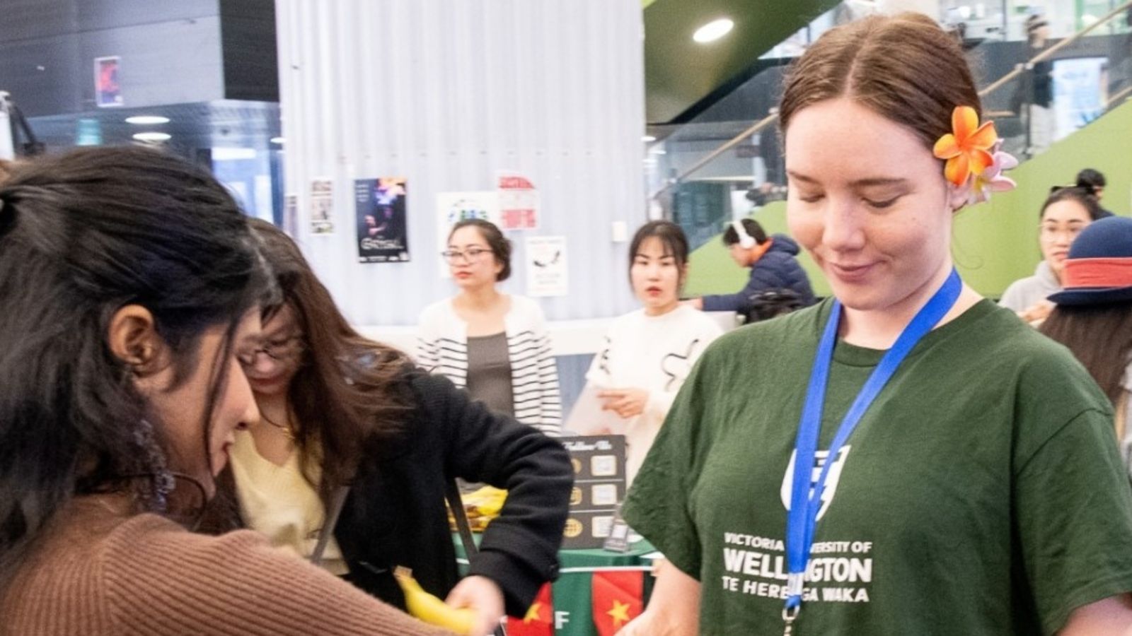 A female Te Herenga Waka staff member is standing in The Hub at Kelburn campus during an event. She is holding a box full of bananas and a female student is about to take some from the box.