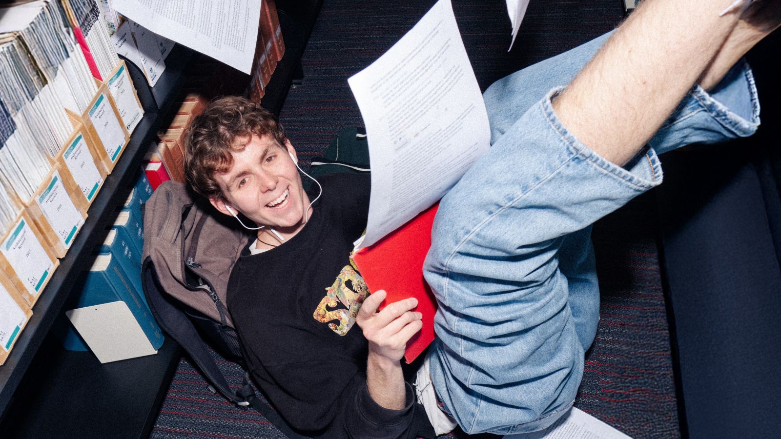 Student lounging on library floor against a backpack. They are wearing earphones and there are papers falling around them