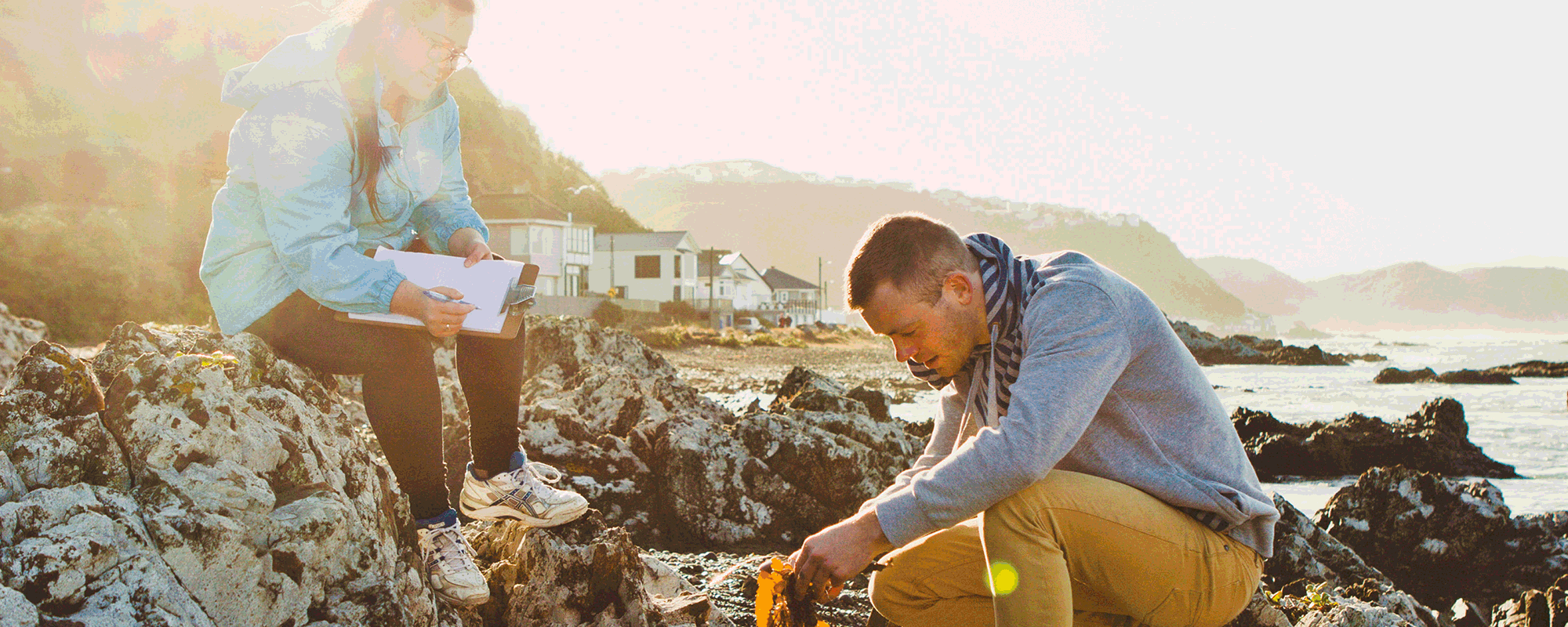 Two research students work are searching through coastal rocks holding a piece of algae.