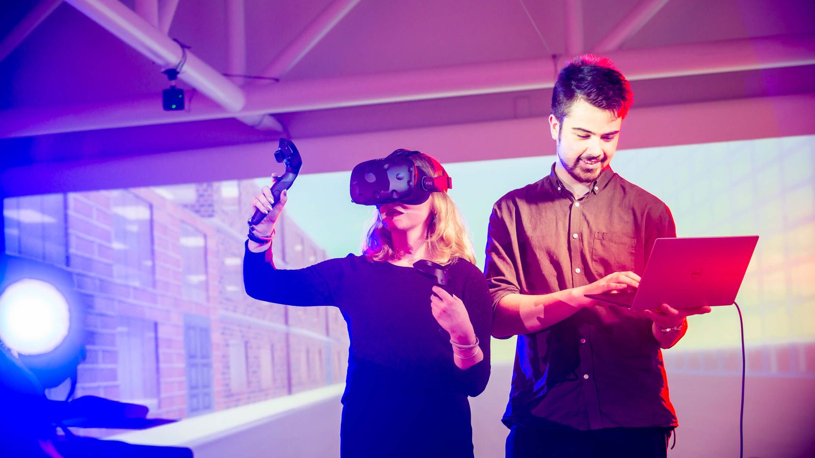 In a room lit by blue and purple lights, a man holds a laptop and types with one hand, and a woman uses virtual reality goggles and a controller.