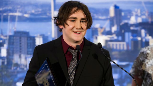Adam Browne stands in a suit at a lectern holding an award with Wellington in the background.