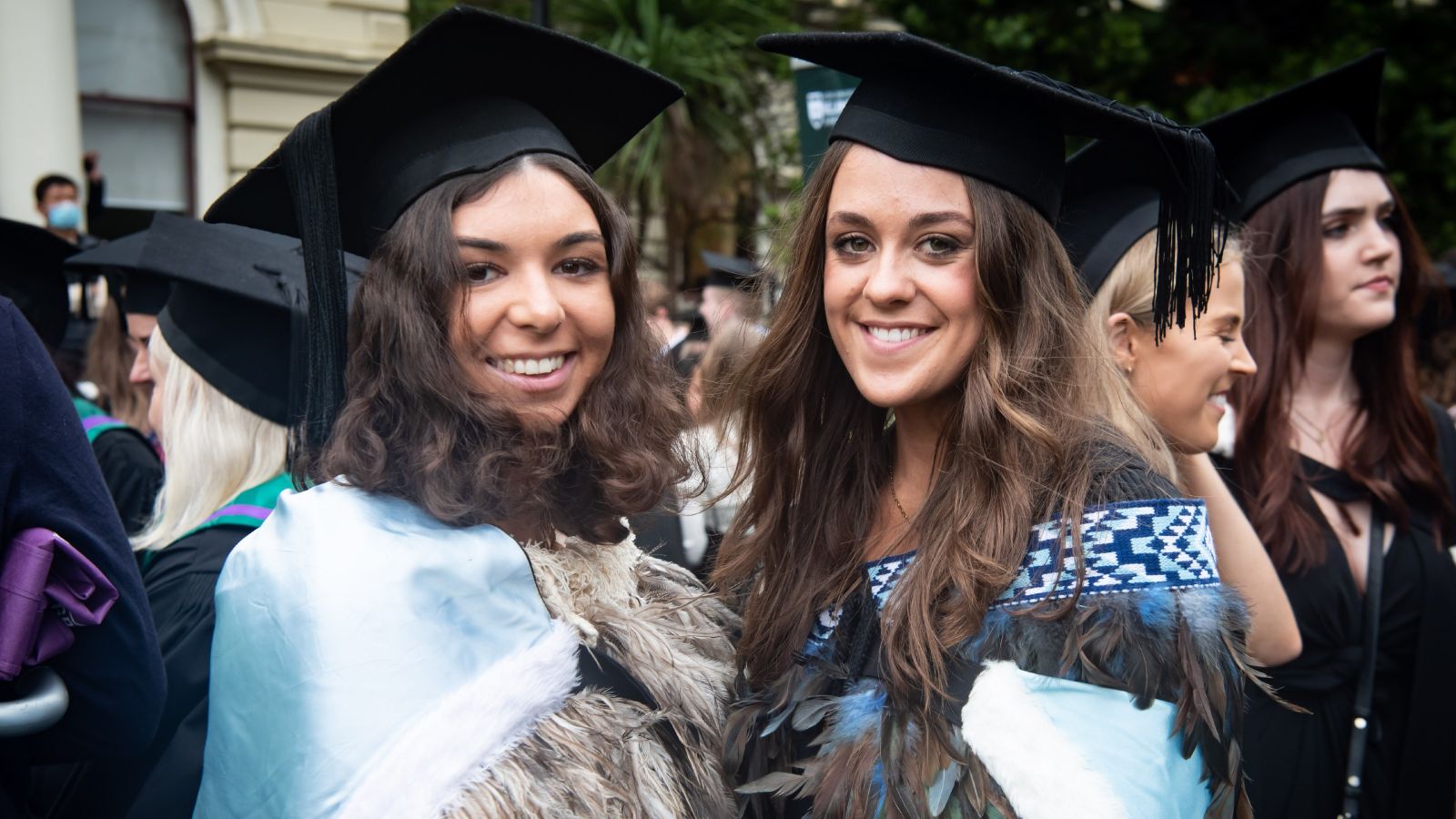 Two young Māori women wearing academic dress and traditional cloaks.