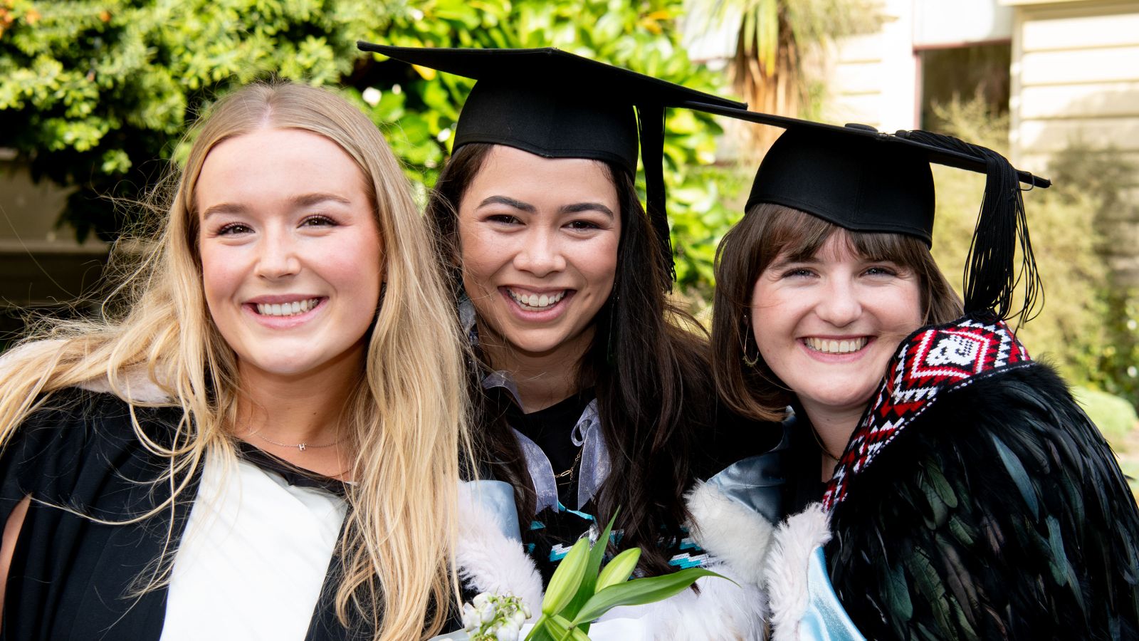 Three students in graduation robes smiling into the camera. Two students are wearing graduation hoods and one is wearing a Korowai 