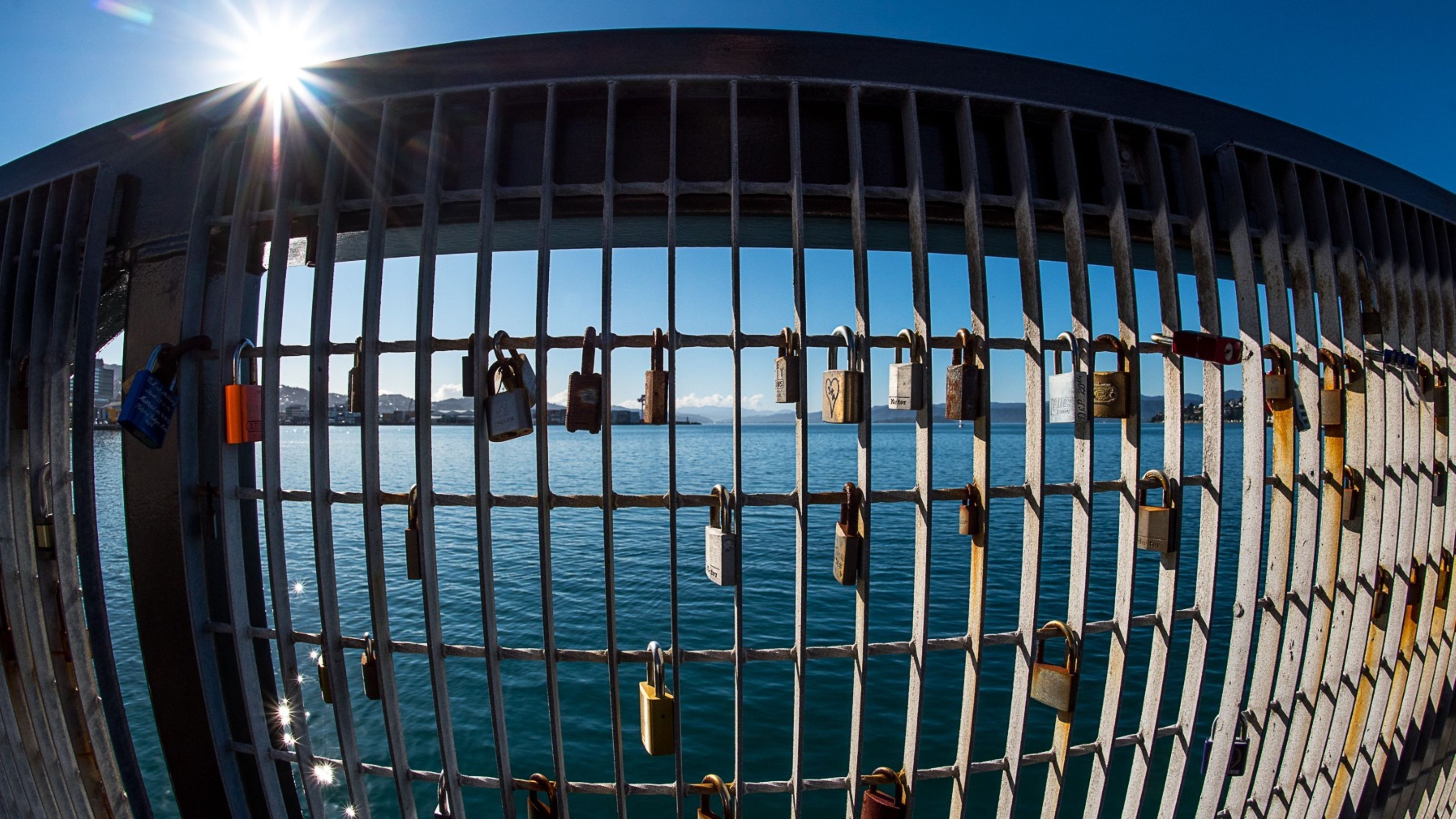 Metal bridge with padlocks attached to the mesh siding with blue sea and sky in the background. 