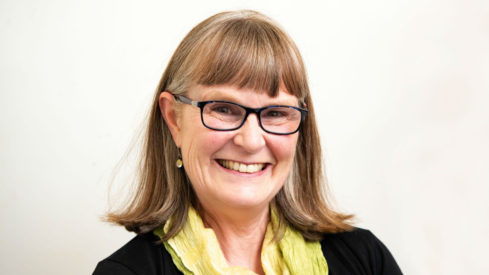 Kay Hancock smiling in a yellow scarf and black rimmed reading glasses. Her hair is blonde, shoulder length, and cut in bangs.