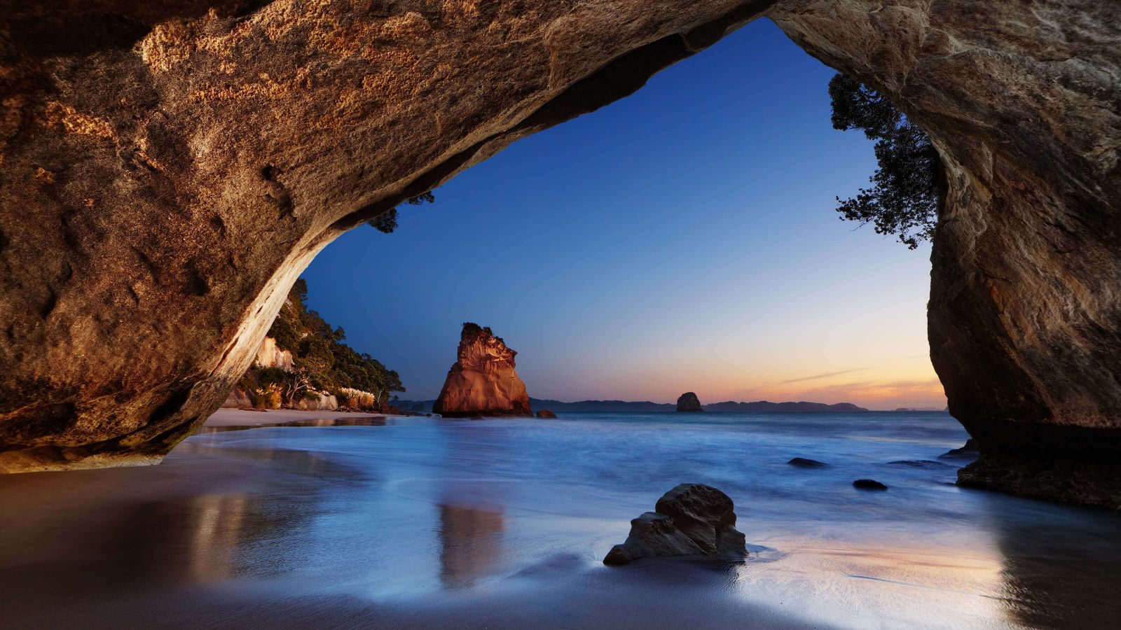 The tide recedes in a twilit seascape framed by the imposing rocky entrance of a huge, seaside cave.