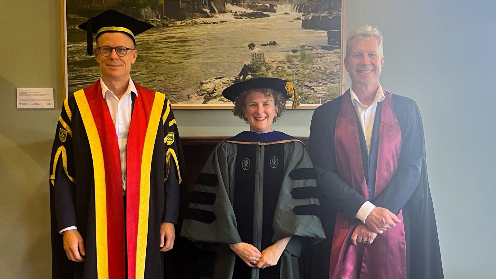 Three professionally dressed academics standing in front of a photo featuring a serene lake scene, with a man engrossed in reading a book by the lakeside.