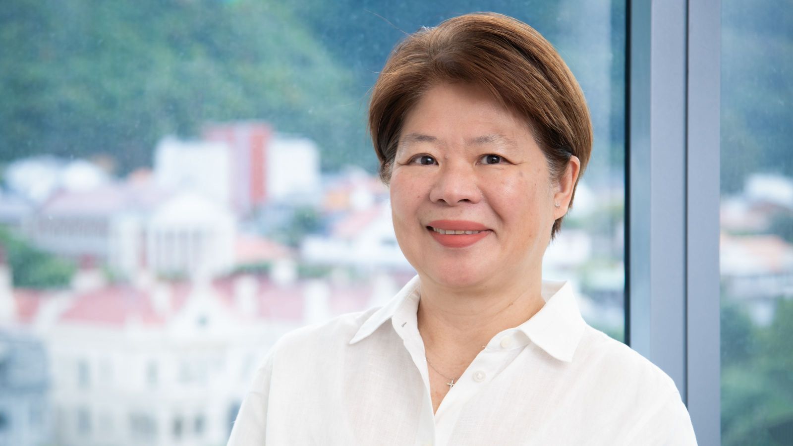 A middle-aged Malaysian woman with copper hair smiling at the camera, wearing a white shirt and with Government Buildings behind her.