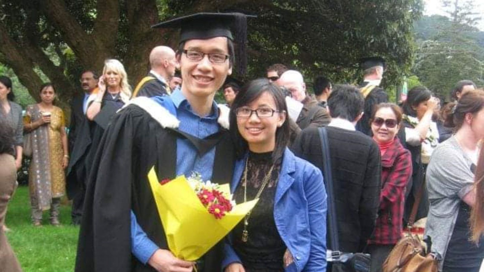 Cong in graduation cap and gown holding flowers and standing with his wife. 