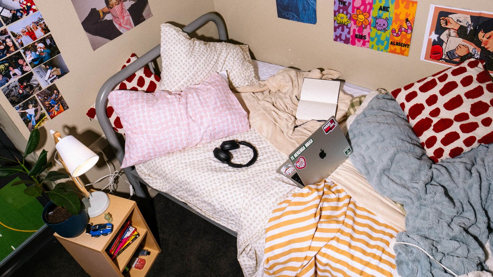 An unmade single bed in a hall dorm. Blankets and pillows are strewn across the bed, along with laptop and earphones. There are posters and photos on the walls